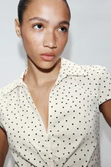 Camisa de cuello solapa y escote en pico, con mangas cortas y detalle de frunces centrales. Presenta cierre frontal con botones y estampado de lunares negros sobre fondo blanco.