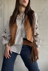 Chaleco de gamuza color camel forrado, con ribetes decorados con tachas metálicas y tiras colgantes con flecos en el lateral.