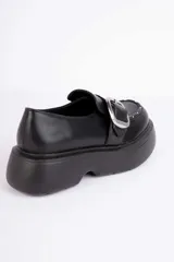 Mocasín negro de plataforma con suela chunky, tira frontal con hebilla metálica plateada y detalles de tachas en el borde del empeine.