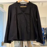 Camisa negra de manga larga, con escote en V con volados sutiles, detalles de encaje y alforzas en el frente, y cierre frontal con botones.