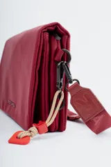 Bandolera de diseño rectangular en color verde oliva, con solapa frontal y logo de la marca grabado. Incluye una correa larga ajustable con el nombre de la marca tejido y un charm decorativo con forma de corazón y letra R en color rojo.