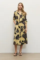 Vestido camisero midi de color amarillo maíz con estampado floral en tonos oscuros, de manga larga, cuello camisero, cierre frontal de botones y cinturón negro delgado con hebilla metálica.
