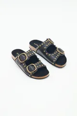 Sandalia plana negra de estilo 'dos hebillas' con tachas metálicas doradas decorando las tiras y hebillas ajustables doradas. La plantilla es de corcho y la suela de EVA.