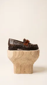 Mocasín de cuero vacuno con textura gastada en tono marrón oscuro. Presenta un detalle de banda frontal en pelo sintético al tono y costuras reforzadas en el empeine.