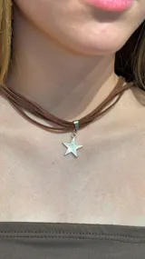 Gargantilla tipo choker compuesta por múltiples tiras finas de gamuza marrón, con un dije central en forma de estrella de metal plateado.