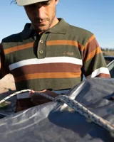 Buzo tejido de punto con diseño de rayas horizontales en tonos verde, marrón y blanco. Presenta cuello tipo polo con cierre de tres botones, mangas largas con puños acanalados y logo de la marca bordado en el pecho. Incluye una etiqueta decorativa en la parte inferior.