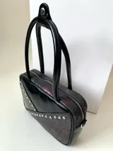 Cartera rígida negra con detalles en rojo y tachas plateadas. Tiene herrajes y cierre metálico plateado marca YKK, asa fina para llevar al hombro o en la mano y correa larga extra regulable. El interior es forrado, tiene un bolsillo con cierre enmarcado en cuero, y un segundo bolsillo plaqué.