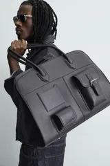Bolso de viaje negro con acabado grabado y diseño de múltiples bolsillos frontales. Dispone de un compartimento principal con cierre magnético, un bolsillo interno con cremallera y tres bolsillos exteriores de diversos tamaños con cierre de imán. Incluye dos asas de mano superiores.