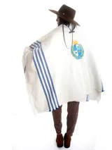 Poncho blanco de hilo de algodón grueso, con franjas azules en los laterales, flecos en el ruedo y el escudo nacional uruguayo bordado en la espalda.