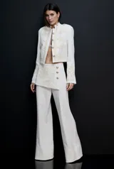 Chaquetilla de sastrería blanca de inspiración militar, con bordados ornamentales claros, cuello mao y botones metálicos dorados en el frente. Presenta hombros estructurados y corte corto.