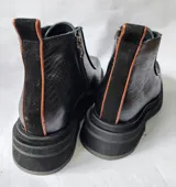 Bota corta de cuero negro con diseño de punta cuadrada y suela track. Presenta un cierre frontal central, detalles de pespuntes y una textura combinada en los laterales.