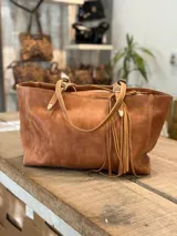 Cartera tipo tote de cuero color tostado, con diseño de doble asa, cierre superior y detalle de flecos laterales.
