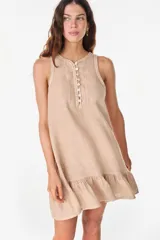 Vestido corto de lino color beige, sin mangas, con escote redondo y cierre frontal parcial con botones de nácar. Presenta un diseño suelto y liviano, con un delicado volado en el ruedo.