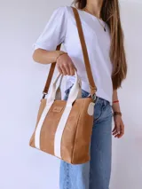 Cartera tipo tote de cuero color camel con detalles de bandas verticales en color crema. Cuenta con doble asa de mano, correa larga ajustable y extraíble, y cierre superior. Incluye etiqueta de cuero con logo grabado en el frente.