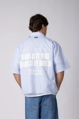 Camisa de manga corta a rayas beige y blancas, de corte boxy y liviana, con cuello camisero, cierre frontal de botones y pequeño texto bordado "BUILD IT BOLD" en el pecho.