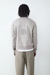 Buzo de cuello redondo y manga larga en color gris claro. Presenta un estampado frontal con tipografía estilo universitario y un diseño gráfico central. Cuenta con puños y cintura acanalados.