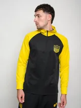 Buzo deportivo de entrenamiento, color negro en el torso y amarillo en las mangas y cuello. Es de manga larga, con cuello alto y cierre parcial frontal. Presenta un escudo con estrellas en el pecho.