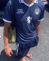 Camiseta de fútbol de Escocia de manga corta, color azul marino con diseño a cuadros sutiles, cuello en V y cuello camisero blanco con líneas azules. Presenta detalles estampados en el frente, incluyendo el escudo de la Asociación de Fútbol de Escocia, un león rampante y el texto 'Scottish Football Association'.