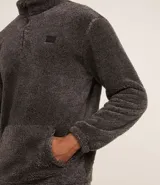 Buzo de tejido peludo color negro, con cuello alto y cierre frontal parcial. Presenta mangas largas con puños elásticos, bolsillo tipo canguro y etiqueta decorativa en el pecho.