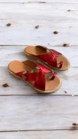 Sandalias planas de cuero rojo metalizado con diseño tipo ojota y detalle de costuras decorativas y aplique metálico lateral. Cuenta con suela de caucho y taco bajo.