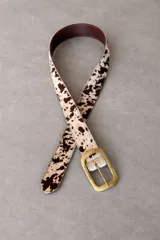 Cinturón ancho de cuero con textura de pelo y estampado animal print de manchas marrones y blancas. Cuenta con hebilla rectangular metálica con terminación envejecida y perforaciones regulables.
