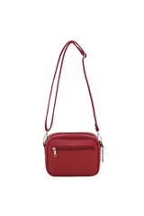 Bandolera color verde oliva con correa larga regulable, cierre principal y bolsillo interno. Tiene una franja horizontal negra en la parte inferior y el logo de la marca en rosa. Incluye un dije colgante en forma de corazón.