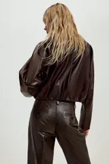 Chaqueta corta de piel color marrón, con cuello redondo, manga larga y cierre frontal con botones. Presenta un diseño con elástico en el ruedo y forro interior.