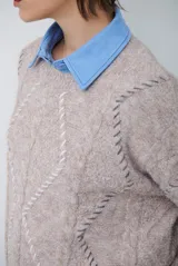 Buzo de punto color beige con diseño de trenzas y costuras decorativas en contraste que forman rombos. Presenta cuello redondo, mangas largas y puños acanalados.