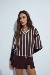 Remera de estilo deportivo con cuello en V y mangas cortas amplias. Presenta un diseño de rayas verticales en tonos marrones, blancos y líneas finas en fucsia, confeccionada en tejido de red.