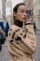 Chaqueta de punto confeccionada en mezcla de lana y alpaca, color beige. Presenta cuello redondo, manga larga, cierre frontal con botones y cinturón ajustable del mismo tejido. Incluye bolsillos delanteros de plastrón y detalles de flores bordadas en color marrón a contraste en la parte superior y en los bolsillos.