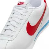Championes Nike modelo Cortez, de diseño clásico en color blanco con el icónico swoosh lateral en rojo. Presenta detalles en rojo en el talón y una franja azul en la entresuela, con suela dentada característica.