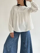 Blusa blanca de corte holgado con cuello redondo, mangas largas con puños elásticos y detalles de puntillas verticales en el frente.