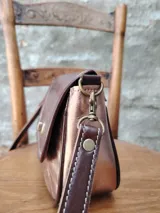 Cartera bandolera pequeña de cuero sintético color bronce con textura craquelada. Tiene correa larga de cuero marrón regulable y cierre metálico dorado.