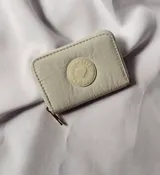 Billetera pequeña de tela impermeable color beige, con cierre perimetral y parche circular central con diseño de corazón y la inscripción Make Up.