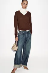 Bolso de mano tipo saca, color borgoña, con asa de mano ajustable y correa bandolera extraíble. Presenta un detalle de doble tira con terminaciones de bolas metálicas doradas en el frente.