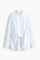 Camisa de algodón blanca, de manga larga con puños abotonados, cuello camisero y cierre frontal de botones. Presenta un diseño con tiras incorporadas en el lateral para anudar, creando un efecto cruzado y drapeado en el frente.