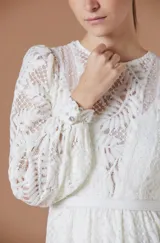 Vestido largo de encaje color blanco, con mangas largas abullonadas, cuello redondo y falda con volados en el ruedo. Presenta un diseño con transparencias y forro interno.