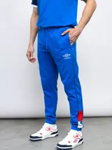 Pantalón de jogging azul de felpa, con detalles de piping y franjas laterales en rojo y blanco. Incluye el escudo del Club Nacional de Football en el muslo derecho.