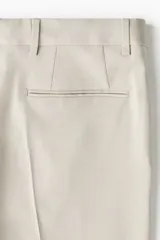 Pantalón de traje beige de corte slim fit, con cierre frontal con cremallera y corchete oculto, trabillas para cinturón, bolsillos laterales y bolsillos traseros ribeteados. Cuenta con raya marcada al frente.