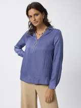 Blusa de satén color azul, con cuello camisero y cierre frontal parcial con cremallera. Presenta mangas largas con puños abotonados y un corte recto y fluido.