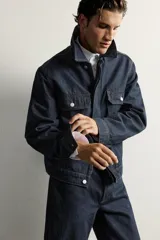 Campera de denim azul oscuro de corte cropped, con cuello camisero, bolsillos delanteros con solapa y cierre frontal oculto con botones metálicos.