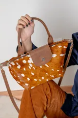 Cartera pequeña de cuero vacuno con diseño de pelo estampado con manchas blancas sobre fondo marrón, tipo animal print. Cuenta con solapa frontal de cuero liso con broche metálico, manija superior, correa larga desmontable y cierre superior.