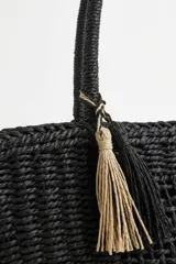 Bolso tipo tote color negro, tejido, con doble asa superior y detalle de borlas colgantes en tonos beige y negro.