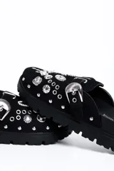 Zueco negro de estilo slip-on, confeccionado con acabado similar a gamuza, suela gruesa dentada de goma, hebilla lateral plateada de diseño occidental y apliques metálicos decorativos con tachas y ojetes en la capellada.