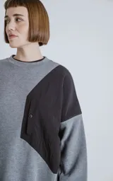 Buzo de cuello redondo con diseño oversize y corte crop. Confeccionado en frisa de algodón color gris melange, presenta un recorte asimétrico en color negro en microfibra con un bolsillo frontal funcional con cierre a presión.