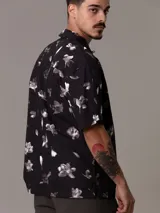 Camisa de manga corta Calvin Klein con corte relaxed fit, confeccionada en algodón regenerativo. Presenta un estampado floral en tonos blanco y gris sobre fondo negro, cierre frontal con botones y un bolsillo de parche en el pecho con el logo de la marca bordado.