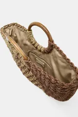 Bolso tipo capazo de rafia tejida, con forma de media luna, en tonos naturales y marrones. Posee un asa rígida de madera en forma de aro y cierre magnético.
