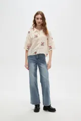 Camisa de algodón color crudo con diseño de paneles unidos por cintas caladas. Presenta bordados florales en cada panel, cuello camisero, cierre frontal con botones y mangas tres cuartos.