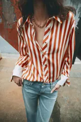 Camisa de manga larga con estampado de rayas verticales en color naranja y crema. Presenta cuello solapa, cierre frontal con botones dorados y puños anchos en color crema con detalle de lazos ajustables.
