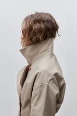 Trench clásico color marrón, de silueta relajada, confeccionado en tejido liviano, con cuello de solapa, doble abotonadura frontal y mangas largas con presillas regulables en los puños.